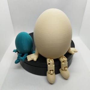 Könnte beinhalten: Eine beige, eiförmige Figur mit beweglichen Beinen sitzt auf einem schwarzen, runden Sockel. Eine kleinere, türkisfarbene, eiförmige Figur mit beweglichen Beinen sitzt neben der größeren Figur.