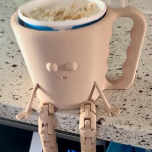 Puede incluir: Una taza caprichosa de color marr&oacute;n claro con una cara, brazos y piernas articuladas. La taza contiene un recipiente blanco lleno de comida. La taza est&aacute; sobre una encimera. Se ve una caja de Rice Krispies.