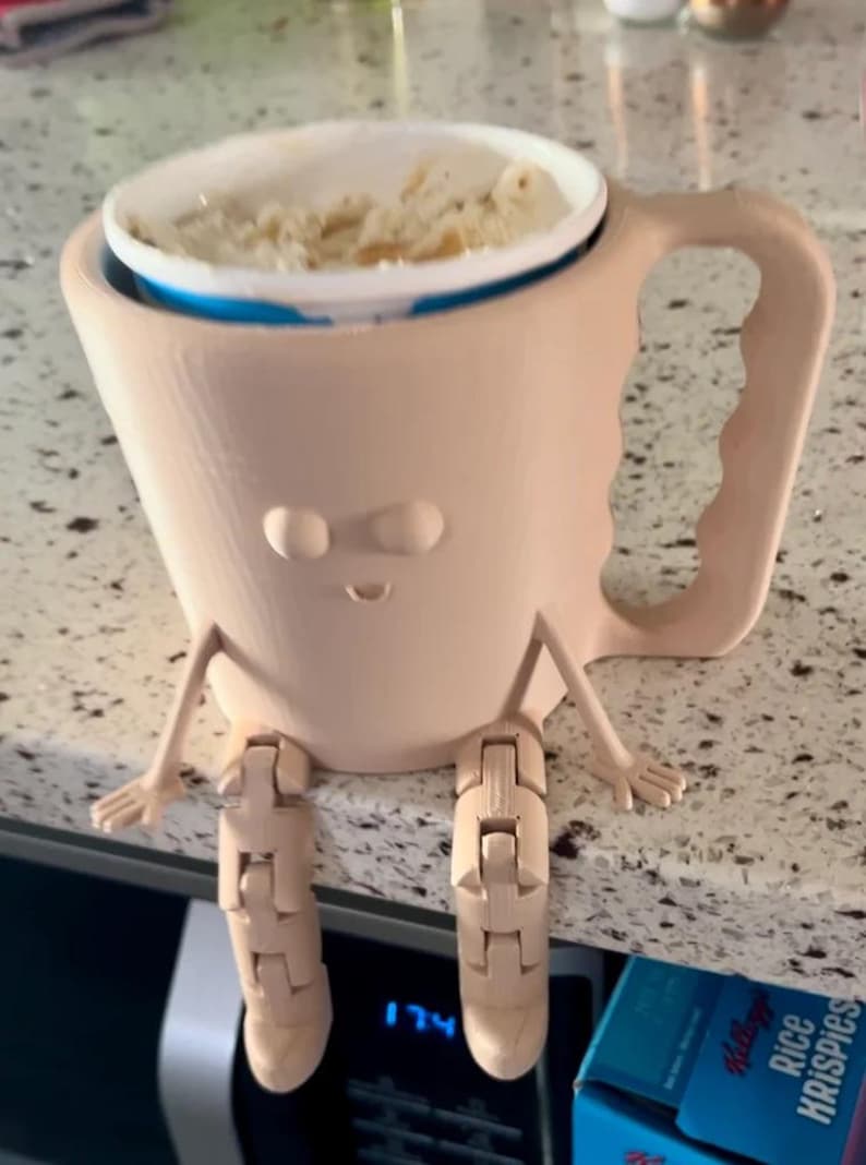 Puede incluir: Una taza caprichosa de color marr&oacute;n claro con una cara, brazos y piernas articuladas. La taza contiene un recipiente blanco lleno de comida. La taza est&aacute; sobre una encimera. Se ve una caja de Rice Krispies.