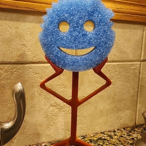 Puede incluir: Una esponja azul con un diseño de cara sonriente, sostenida por un soporte geométrico rojo. La esponja tiene dos agujeros para los ojos y una boca curvada. El soporte es de plástico rojo y tiene una base cuadrada.
