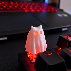 May include: A white, ghost-shaped mechanical keyboard keycap featuring two circular eye cutouts. The keycap is mounted on a black keyboard switch that emits a bright red light, which illuminates the interior of the ghost figure.