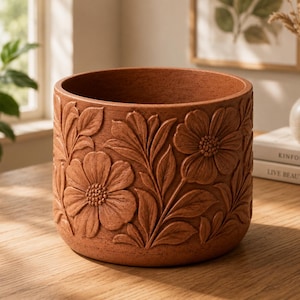 May include: A cylindrical terracotta-colored planter featuring a textured surface with an embossed floral pattern of flowers and leaves wrapping around the exterior. The pot has a wide circular opening and a flat base, showcasing a matte finish.