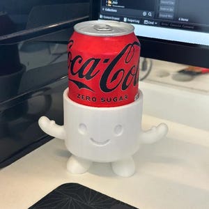 May include: A red Coca-Cola Zero Sugar can is held by a white, smiling, marshmallow-shaped holder with arms and feet. The can features a black script logo.