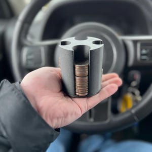 May include: A matte black cylindrical coin organizer featuring four vertical slots designed to hold stacks of coins. The device has a textured finish and is shaped with a cross-like top opening, allowing for the organized storage and retrieval of currency.