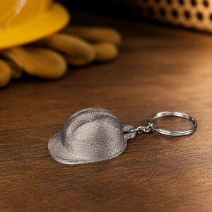 Könnte beinhalten: Ein silberfarbener Metallschlüsselanhänger in Form eines Miniatur-Bauhelms. Der Schlüsselanhänger verfügt über einen standardmäßigen runden Metallring, der an der Rückseite des kleinen, texturierten Schutzhelms befestigt ist und eine realistische Form aufweist.