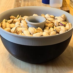 May include: A two-tone snack bowl filled with pistachios. The bowl is grey and black with a central compartment. The pistachios are in their shells, with a light tan colour. The bowl sits on a wooden surface.