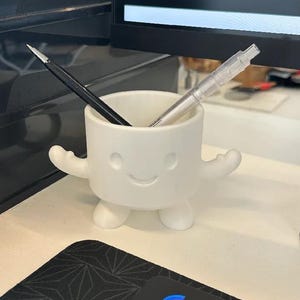 Puede incluir: Un organizador de escritorio de cer&aacute;mica blanca con forma de personaje sonriente con brazos y piernas. Un l&aacute;piz negro y un portaminas plateado est&aacute;n dentro. El organizador est&aacute; sobre una superficie de escritorio blanca.