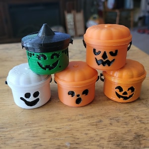 May include: A collection of small, 3D-printed Halloween-themed containers. The containers are shaped like pumpkins, a witch's cauldron, and a ghost. They are orange, green, white, and black, with carved-out faces. The containers are stacked on a wooden surface.