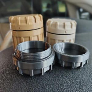 Puede incluir: Se muestran tres contenedores impresos en 3D. Dos son de color beige y blanquecino, con una parte superior estriada y un cuerpo cilíndrico. Los otros dos son de color gris oscuro, con un interior roscado y un exterior estriado. Los objetos están sobre una superficie oscura.