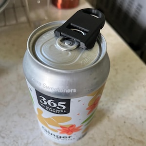 Può includere: Un apri-lattine nero è posto sopra una lattina di Ginger Sparkling Water di 365 Whole Foods Market. La lattina ha uno sfondo bianco con disegni floreali arancioni e la scritta "Zero Sweeteners".