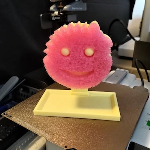 Puede incluir: Una figura impresa en 3D de una cara sonriente rosa y amarilla. La figura tiene una apariencia texturizada, similar a una esponja, con dos ojos circulares blancos y una boca sonriente. Está montada sobre una base rectangular amarilla.