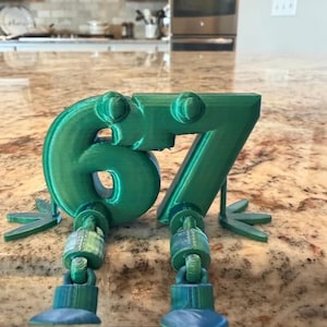 May include: A green 3D-printed number 67 with articulated legs and feet. The digits are in a playful font, with the legs and feet attached to the bottom of the numbers. The feet are a lighter shade of blue.