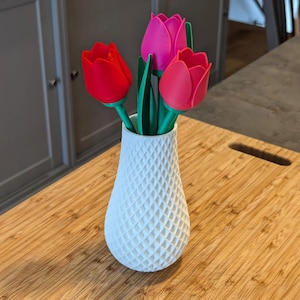 May include: A white, textured vase holding three artificial tulips. The tulips are red and pink, with green stems and leaves. The vase is on a wooden cutting board.