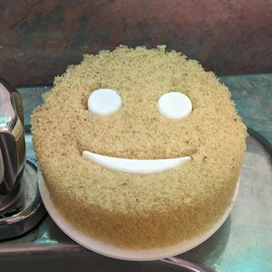 May include: A round, tan sponge with a smiling face design. The face is created with two white, circular eyes and a white, curved mouth. The sponge sits on a white, circular base, likely for use in a kitchen or bathroom.