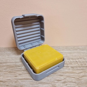 May include: A grey, square soap dish with a hinged lid, open to reveal a yellow bar of soap. The dish has a ridged interior for drainage. The soap is rectangular and fills the dish. The background is a light brown.