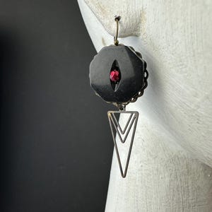 May include: A dangle earring featuring a matte black octagonal component with a central vertical slit revealing a small, faceted red bead. Below the octagon hangs a geometric, open-frame triangular pendant made of dark, antiqued metal wire.