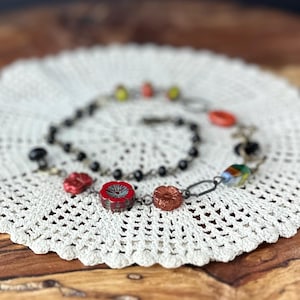 May include: A multi-strand necklace featuring a mix of black beads, metallic red flower-shaped beads, orange oval beads, and a multi-coloured glass bead. The necklace is constructed with dark metal chain links and includes a lobster claw clasp closure.