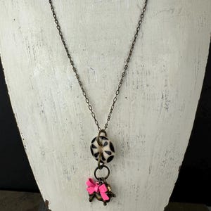 Puede incluir: Un collar largo de cadena marrón con una cuenta de patrón blanco y negro y un dije de lazo rosa.