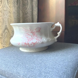 May include: White porcelain chamber pot with a pink floral design. The antique pot has a handle and a pedestal base. The floral pattern is a delicate rose motif. The pot is sitting on a gray upholstered surface.