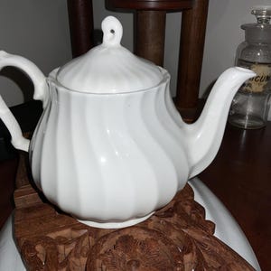May include: White ceramic teapot with a lid and a curved handle. The teapot has a spiral design and a spout. The teapot is sitting on a wooden stand. A glass bottle and a white pitcher are in the background.