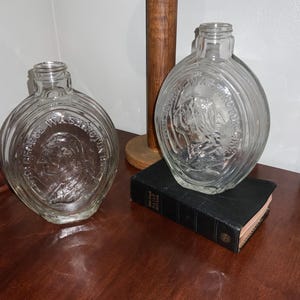 May include: Two clear glass flasks with oval bodies and embossed portraits of George Washington. The flasks are displayed on a dark wooden surface. One flask rests on a small, black book.