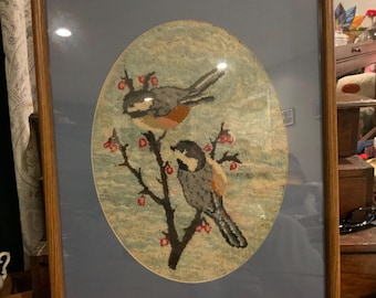 May include: Framed artwork featuring two birds perched on a branch with red berries. The birds are depicted in shades of gray, brown, and orange against a light blue and green background. The artwork is encased in a wooden frame.