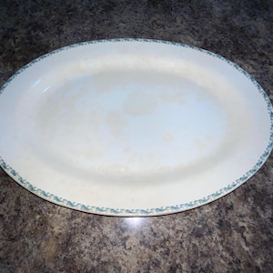 May include: An oval-shaped serving platter with a cream-colored center and a decorative teal green border pattern along the rim. The surface of the platter shows subtle signs of age and discoloration, featuring a smooth, glossy finish throughout.