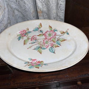May include: Oval, off-white ceramic platter with a floral design. The flowers are pink with green and blue leaves. The platter has a gold rim and is likely vintage or antique.