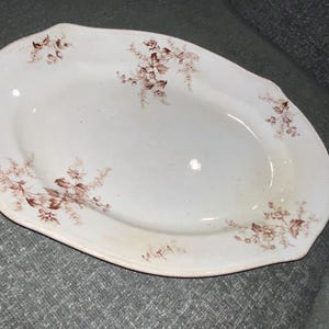 May include: An oval-shaped white ceramic serving platter featuring a delicate brown floral pattern printed along the scalloped edges. The surface is smooth and glossy, with the botanical illustrations distributed across the rim of the dish.