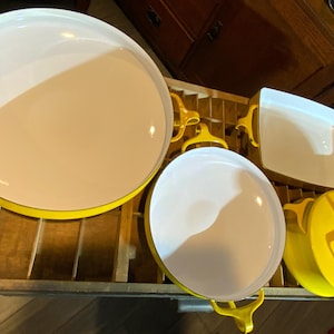 May include: A collection of white and yellow enamelware cookware. The set includes a large round tray, a smaller round pan, a rectangular baking dish, and a round lid. The cookware has a white interior and a yellow exterior with yellow handles.