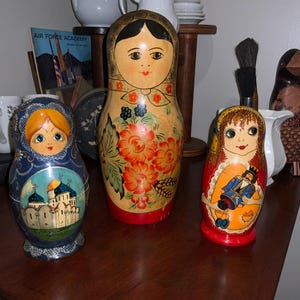 May include: Three Russian nesting dolls, or Matryoshka dolls, stand on a wooden surface. The largest doll is beige with floral and facial details. The other two dolls are smaller, one blue with a church scene, and the other red with a figure.