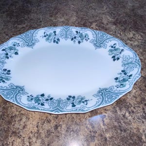 May include: An oval-shaped white ceramic serving platter featuring a scalloped edge and a decorative border pattern in teal green. The border consists of intricate scrollwork and floral motifs that frame the plain white center of the dish.