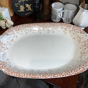 May include: An oval, white ceramic serving platter with a brown floral pattern around the rim. The platter has two handles and is surrounded by other decorative items, including mugs and a small rabbit figurine.