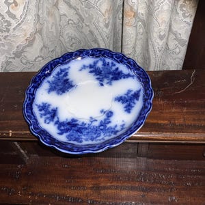 May include: A small, antique blue and white saucer with a scalloped edge. The saucer features a floral pattern in shades of blue against a white background. The dish is likely made of porcelain and is displayed on a dark wooden surface.