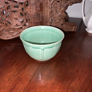 May include: A vintage, light green ceramic bowl with a scalloped rim. The bowl has a simple, elegant design and is set on a dark wooden surface. The bowl is empty and appears to be in good condition.