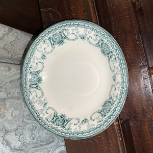 May include: White ceramic bowl with a teal floral design around the rim. The bowl has a wide, shallow shape and is likely vintage. The teal pattern includes swirls and floral motifs, adding a decorative touch to the piece.
