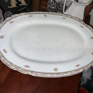 May include: An oval, white porcelain serving platter with a scalloped edge. The rim features a delicate floral and gold trim design. Small pink rose clusters are evenly spaced around the edge. A classic piece for serving food.