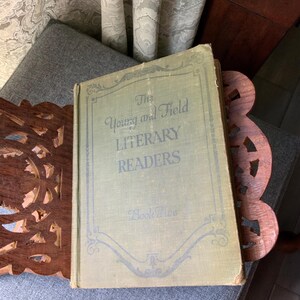 May include: An antique book titled "The Young and Field Literary Readers" with a light green cover and ornate lettering. The book is resting on a wooden book stand with a decorative cutout design. The background includes a gray cushion and patterned fabric.