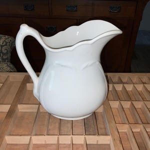 May include: A white ceramic pitcher with a curved handle and a scalloped rim. The pitcher has a bulbous body and a decorative floral design. It is sitting on a wooden surface.