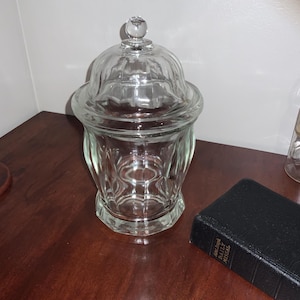 May include: A clear glass jar with a lid, featuring a decorative design. The jar is on a wooden surface, alongside a small book and a glass bottle. The jar's lid has a small, round knob.