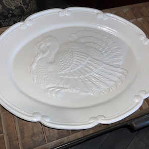 May include: An oval-shaped white ceramic serving platter featuring a raised, embossed turkey design in the center. The platter has a smooth, glossy finish and a decorative scalloped edge with rounded detailing along the entire outer rim.