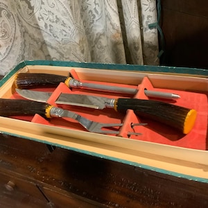 May include: A vintage carving set in a box. The set includes a carving knife, fork, and sharpening steel, all with brown and yellow handles. The set is displayed in a box with a red interior.