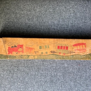 May include: A vintage wooden pencil box featuring a hand-painted train scene. The box is rectangular, with a light brown background and a green base. The train cars are red, green, and orange, with white clouds above.