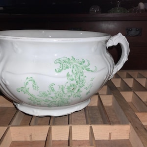 May include: A white ceramic chamber pot with a decorative green floral design. The pot has a rounded shape, a wide rim, and a handle. The pot is sitting on a wooden surface.