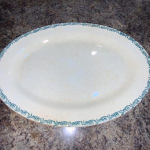 May include: An oval-shaped cream-colored ceramic serving platter featuring a decorative teal-colored border pattern along the rim. The platter has a smooth, glossy finish and a wide, shallow basin, providing a flat surface for food presentation.