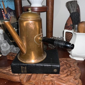 May include: A vintage copper coffee pot with a black handle rests on a black book. The pot has a spout and a lid. A white pitcher with brushes and other items is in the background. A book with the words "Air Force Academy" is also visible.
