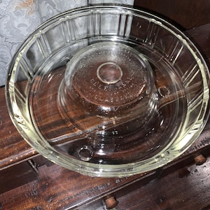 May include: Clear glass serving dish with a central, raised section and a wide, shallow outer ring. The dish has a ribbed design on the outer edge and the text "CLEAN GLASS" is visible on the central section. The dish is sitting on a dark wooden surface.