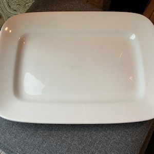 May include: A rectangular, white ceramic serving platter with rounded corners. The dish has a smooth, glossy surface and is designed for serving food. The platter is a simple, elegant design.