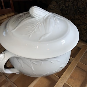 May include: A white ceramic serving dish with a lid, featuring embossed corn and leaf designs. The dish has a handle and is likely used for serving food. The overall design is elegant and detailed, suitable for a formal dining setting.