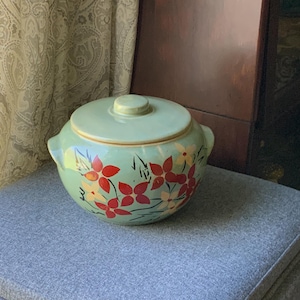 May include: A vintage, light green ceramic casserole dish with a matching lid. The dish is adorned with red and yellow floral designs. It has two handles and sits on a gray upholstered ottoman.